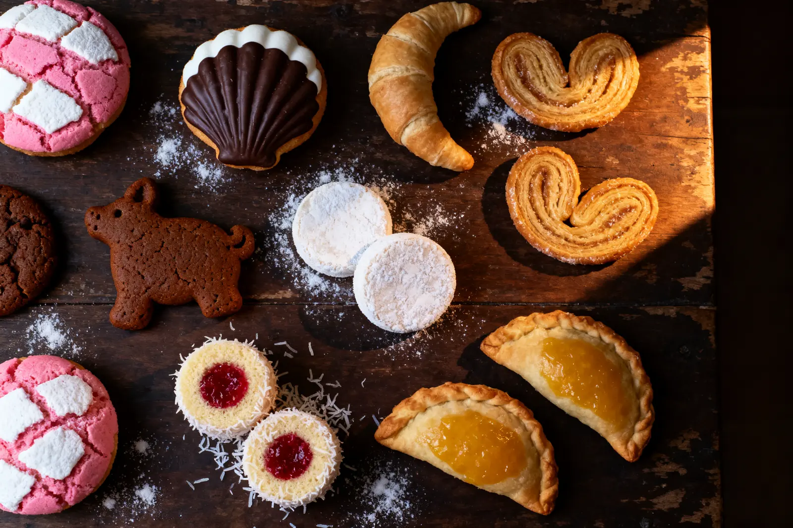 Assorted pan dulce on a dark wooden table: conchas, orejas, a cuerno, a marranito, polvorones, yoyos and empanadas